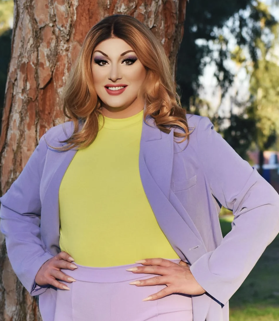 You see a photo of Maebe A Girl, Standing proud with both fiercely-manicured hands on her hips.  Red shoulder length hair, full make up with red lips, a lavender pantsuit with a  yellow top underneath. She's standing in front a a large tree in a park setting.